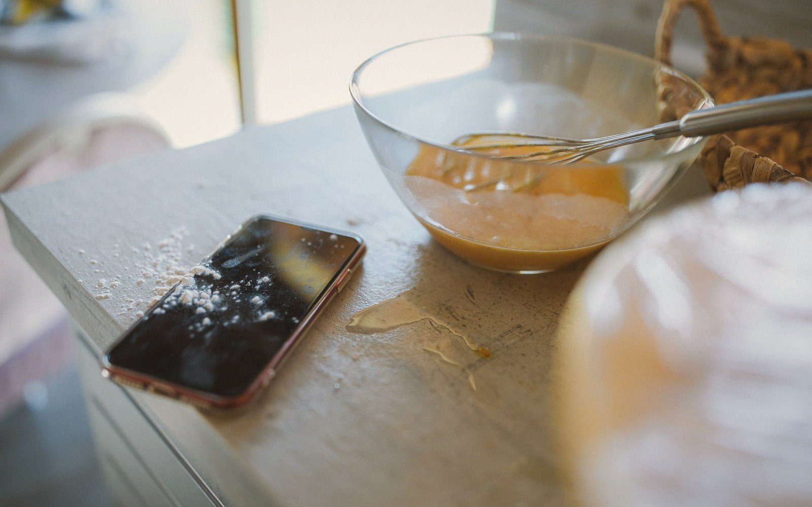 Whisk in a bowl of batter beside a flour-dusted phone on a counter