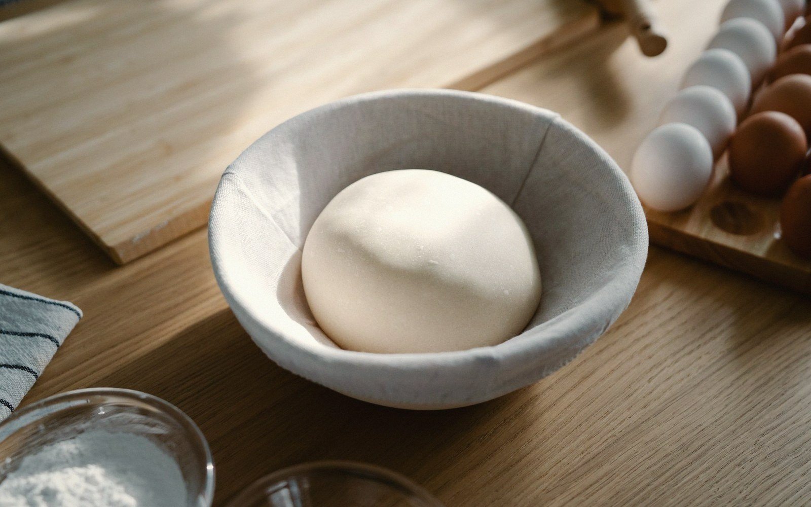 Ball of dough resting in a cloth-lined bowl beside eggs and a bowl of flour