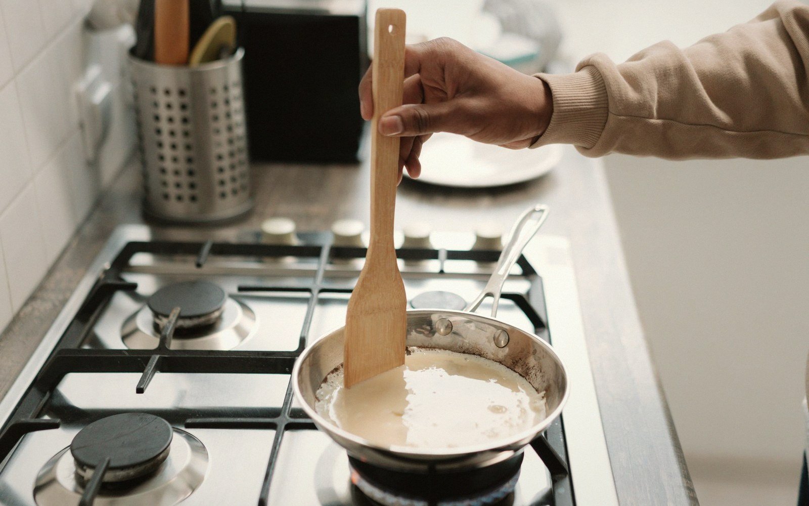 A hand stirs a wooden spatula in milk simmering in a pan on a gas stove